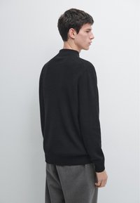 Jeune homme de profil portant un pull en maille noire et un pantalon gris sur un fond clair uni.