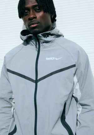 Pilkas sportinis džemperis su užtrauktuku priekinėje dalyje, su juodais akcentais, gobtuvu ir ryškiu "BARCA" logotipu. Lygus paviršius, ergonomiškas dizainas ir šoniniai kišeniai.