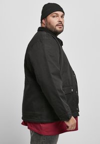 Giacca nera in tela con chiusura a bottoni e tasche laterali, indossata sopra una maglietta rossa. Il modello posiziona di lato, indossando un berretto a maglia nero.