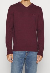 Burgundowy sweter z dekoltem w kształcie litery V, wykonany z miękkiej dzianiny, z długimi rękawami i ściągaczami, ozdobiony małym logo na piersi.