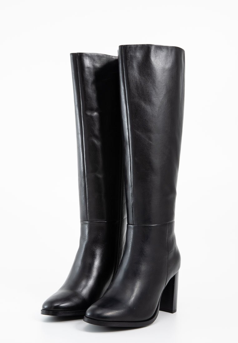 Para czarnych, skórzanych botków do kolan z wysokimi obcasami w kształcie bloku i spiczastymi noskami, stojących prosto na białym tle.