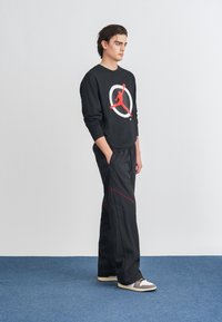 Μαύρη φούτερ μπλούζα με κόκκινο γραφικό και λογότυπο, σε συνδυασμό με μαύρο παντελόνι με κόκκινες ραφές. Το μοντέλο στέκεται σε μπλε χαλί, φορώντας γκρι αθλητικά παπούτσια.