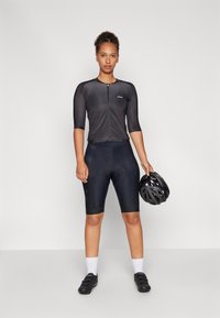 Schwarzes Radtrikot mit kurzen Ärmeln und Reißverschluss, kombiniert mit schwarzen gepolsterten Shorts. Das Modell hält einen schwarzen Helm in einer Hand.