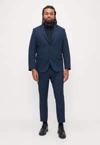 Dunkelblaues Sakko und Hose, mit einem schwarzen Hemd zum Knöpfen. Glatter Stoff, tailliertes Design, zwei Fronttaschen und glänzende schwarze Schuhe.