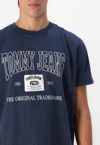 Jūrinės medvilnės marškinėliai su baltais užrašais: "TOMMY JEANS", "ATHLETIC DIVISION" ir "THE ORIGINAL TRADEMARK". Apvalus iškirpimas ir trumpomis rankovėmis.