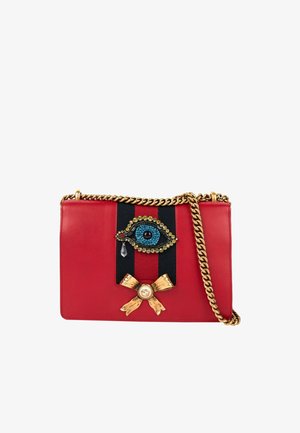 Bolso de cuero rojo para llevar al hombro con cadena dorada, franja negra y roja, parche de ojo con joyas y lágrima, y detalle de lazo dorado.