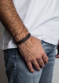 Viekst reimēts ādas aproce tumši brūnā krāsā, ar gludu tekstūru un noapaļotu formu, uz plaukstas, kas ir tērpta ikdienišķā džinsa un baltajā krekliņā.