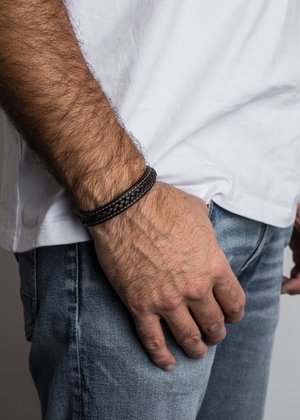 Austere brūns ādas pinumu rokassprādze ar gludu tekstūru un apaļu formu, valkāta uz rokas ar ikdienišķiem dīna auduma izstrādājumiem un baltu krekli.