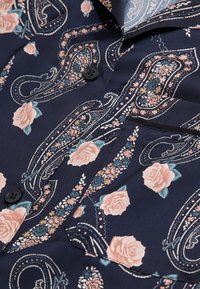 Camicia navy con bottoni, decorata con motivi paisley e floreali in rosa chiaro e teal. Tessuto morbido con una tasca e bottone nero a contrasto.