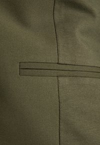 Tissu vert olive à texture lisse, avec une couture piquée et une seule poche verticale. Lignes épurées et design minimaliste.