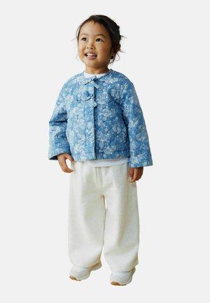 Blå quiltet jakke med blomsterprint og knaplukning, hvid skjorte med lange ærmer og løse cremefarvede bukser, kombineret med lyse sneakers.