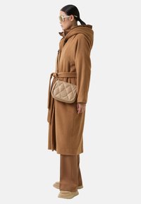 Brun uldblandingsfrakke med hætte, fastgjort talje, båret over brune bukser og beige fodtøj; cremefarvet quiltet crossbody taske med logo detalje.