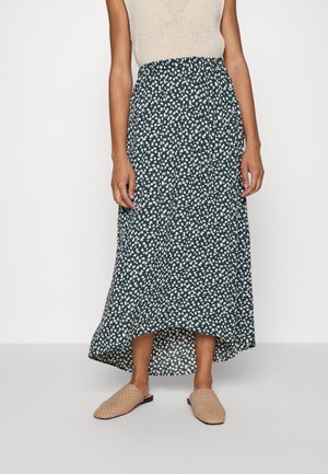 Femme portant une jupe midi vert foncé à petits motifs de feuilles blanches, un haut beige sans manches et des chaussures beige tissées sans lacets.