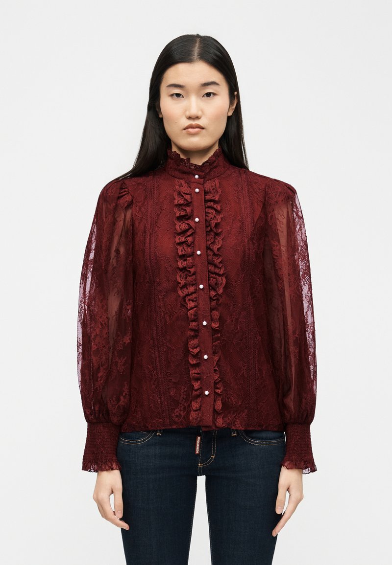 Γυναίκα με μακριά σκοτεινά μαλλιά φοράει μπορντό δαντελωτή μπλούζα με βολάν, κουμπιά από πέρλες, φουσκωτά λεπτά μανίκια και σκούρο μπλε τζιν.