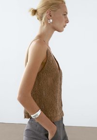 Brown lace tank top with a deep V-neck, textured surface, and small embellishments, paired with gray pants and a silver bracelet.