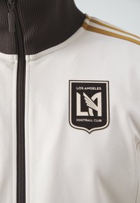 Bílá sportovní bunda na zip s černým zipem a hnědým vroubkovaným límcem. Obsahuje logo LAFC v černobílé barvě a zlaté pruhy na ramenou.