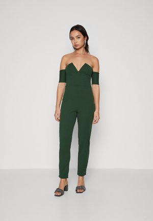 Πράσινο, εφαρμοστό ολόσωμο σαλόνι με βαθύ ντεκολτέ και μανίκια που πέφτουν στους ώμους, σε συνδυασμό με μαύρες ψηλοτάκουνες σάνταλες. Λείο ύφασμα.