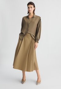 Blusa verde oliva con dettagli di bottoni sulle maniche, abbinata a una gonna fluida coordinata. Tessuto testurizzato con una finitura liscia, indossata con tacchi a fantasia.
