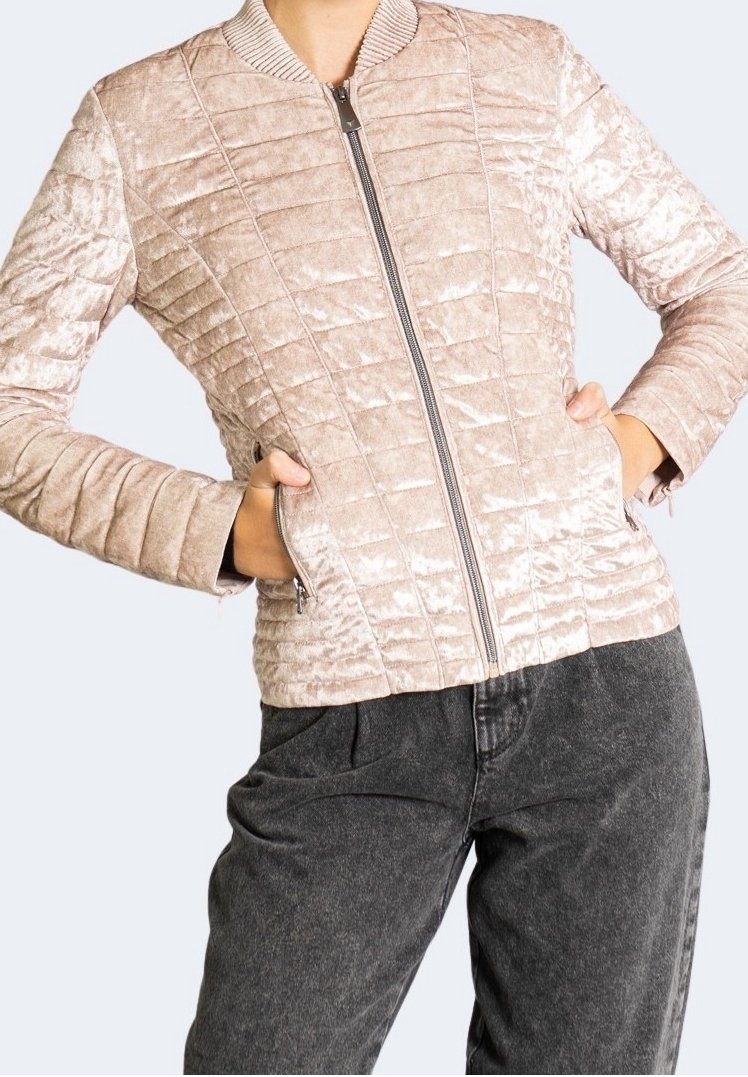 Veste matelassée en velours beige avec fermeture éclair sur le devant, col côtelé et poches latérales zippées, portée avec un pantalon gris foncé.