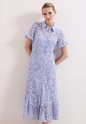 Frau trägt ein hellblaues Spitzen-Midikleid mit kurzen Ärmeln, Kragen, vorderen Knöpfen und Rüschensaum und steht vor einer schlichten Wand.