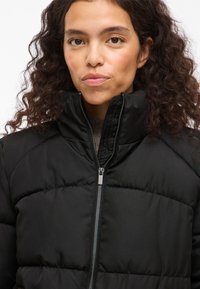 Čierna prešívaná bunda s prešívaným dizajnom, vysokým golierom a striebornými detailmi na zips. Hladká látka s matným povrchom.