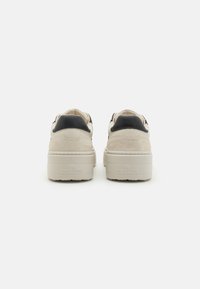 Baskets en daim beige avec des accents en cuir noir, présentant une semelle blanche épaisse. La vue arrière montre le logo embossé sur le talon.