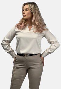 Bluzka w kolorze kremowym, z długim rękawem, z dekoltem w kształcie litery V i szczegółami w formie dziurki, zestawiona z beżowymi spodniami i czarnym paskiem z metalową klamrą.
