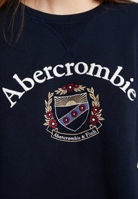 Sudadera navy con la palabra "Abercrombie" en letras blancas y un emblema bordado circular con flores y un diseño de escudo.