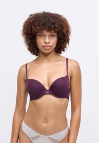 Kvinne med krøllete hår iført en lilla underwire-bh og lyse beige mesh-truser mot en enkel hvit bakgrunn.
