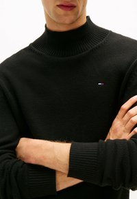 Jersey de cuello alto negro con textura acanalada. Presenta un pequeño logotipo tricolor en el pecho y puños acanalados para un detalle adicional.