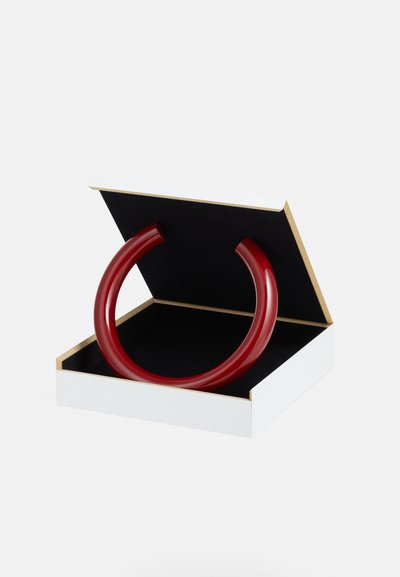 Pulsera de acrílico rojo exhibida en una caja blanca abierta con un interior negro. La caja presenta un acento dorado en el borde.