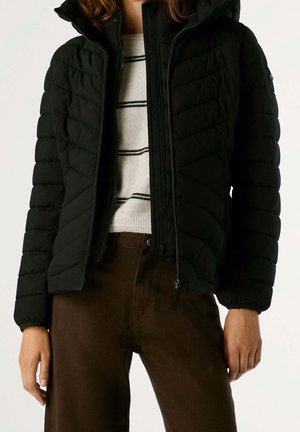 Femme portant une veste matelassée noire avec capuche, un pull blanc avec des rayures noires et un pantalon marron, debout devant un fond uni.