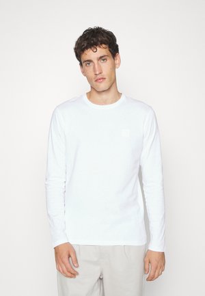 Camisa blanca de manga larga hecha de algodón con un cuello redondo. Presenta un pequeño parche cuadrado en el pecho. Textura suave, diseño simple.