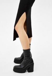 Bottes à plateforme noires avec extérieur en cuir lisse, design ample à la cheville, talon épais et coutures détaillées. Associées à une robe noire fendue.