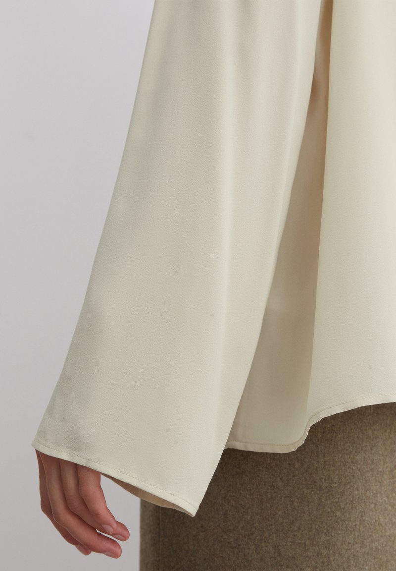 Langärmlige, cremefarbene Bluse mit glatter Textur und weiten Ärmeln. Der untere Saum ist leicht gewölbt und wird mit einem braunen, strukturierten Rock kombiniert.