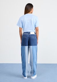 Svetlo modra majica s kratkimi rokavi z belim logotipom Adidas; v kombinaciji z večbarvnimi jeans hlačami iz različnih odtenkov modre.
