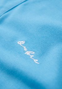 Světle modrý textil s texturovaným povrchem, se bílou vyšitou logem v dolním pravém rohu.