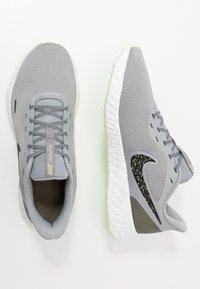 Baskets de sport gris clair avec un dessus en mesh texturé, un swoosh accentué de noir et de vert, et une semelle blanche rembourrée. Lacets et col rembourré.