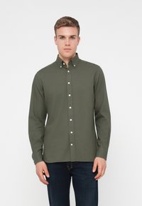 Camisa de manga larga color verde oliva con cuello abotonado, botones blancos y un logo sutil en el pecho. Tejido suave, corte entallado.