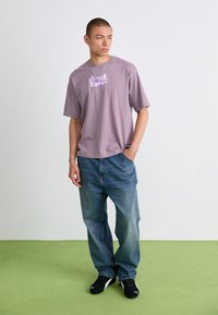 Ung mand står på grøn gulv iført en lavendel grafisk t-shirt, løse blå jeans, sorte sneakers og en sølv kædehalssmykke.