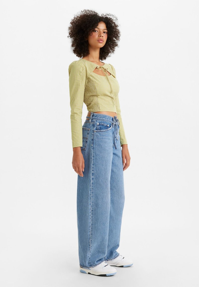 Helles grünes, tailliertes Langarmoberteil mit einer gebundenen Schlüsselloch-Details, kombiniert mit lockeren hellblauen Denim-Jeans. Weiße Sneakers vervollständigen das Outfit.