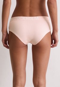 Kobieta w jasnoróżowych majtkach z szerokim pasem, na którym widnieje delikatny napis "STOCKHOLM", prezentowana od tyłu na jednolitym tle.