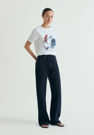 Femeie stând cu mâinile la spate, purtând un tricou alb cu grafică, blugi largi închiși la culoare cu șnur și sandale negre pe fundal simplu.