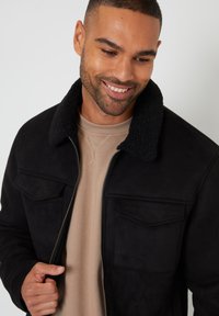 Sort ruskindjakke med en shearling-foret krave, der har en frontlynlås og to brystlommer. Båret over en beige crewneck trøje.
