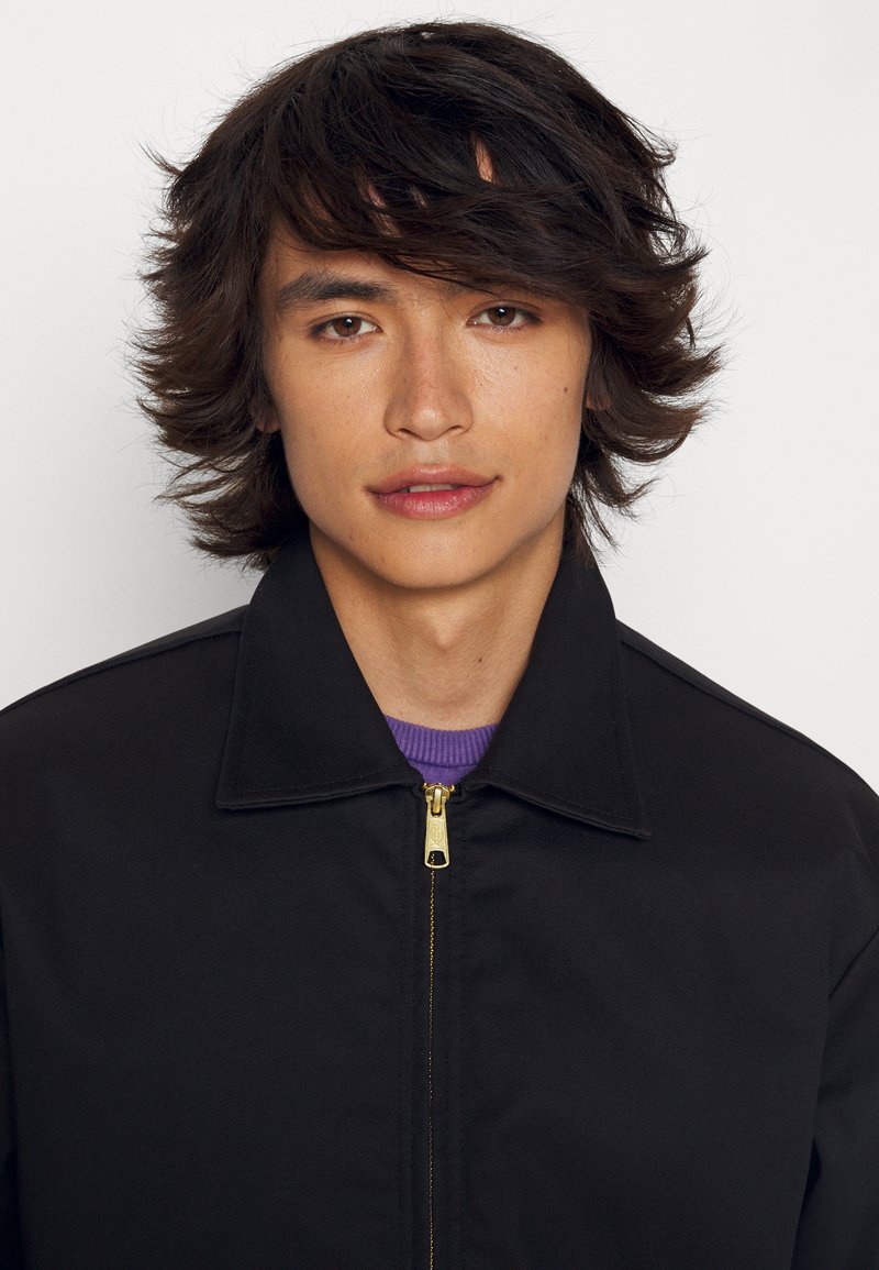 Chaqueta negra con cuello y cremallera dorada, superpuesta sobre una camisa morada. La tela tiene una textura suave y un corte limpio.