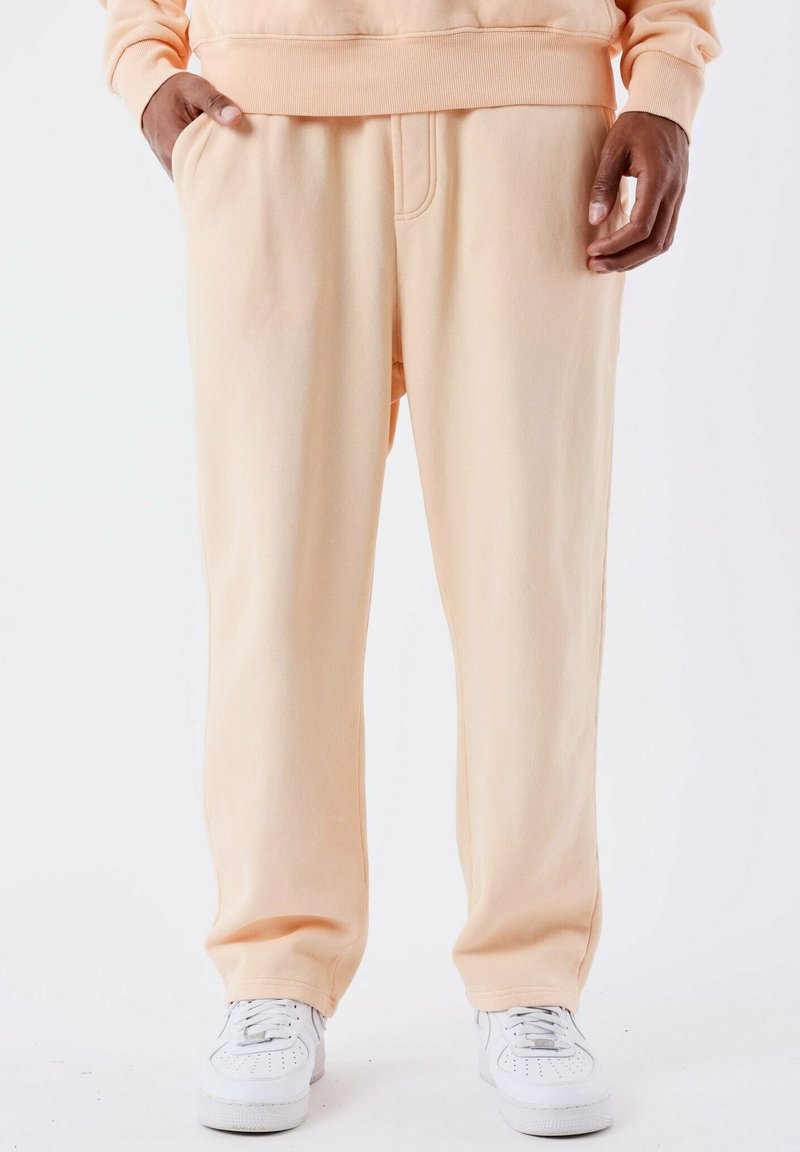 Pantalones de chándal de un suave color melocotón claro, confeccionados en material suave, que cuentan con una cinturilla elástica, bolsillos laterales y un corte relajado. Se combinan con zapatillas blancas.