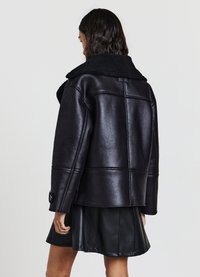 Giacca nera in ecopelle con un colletto peloso e testurizzato e dettagli con bottoni; presenta una forma strutturata e cuciture visibili.