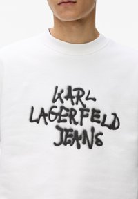 Bijela dukserica s teksturiranim materijalom i crnim logotipom spraya koji glasi "KARL LAGERFELD JEANS." Jednostavan dizajn s okruglim izrezom.