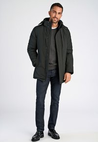 Mörkgrön puffjacka med huva, med blixtlåsfickor och rak passform. Buren över en grå kabelstickad tröja och mörka jeans.