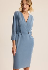 Niebieska sukienka-wrap z rękawami o długości trzech kwart, elastyczną talią i detalem guzika. Gładka faktura i dopasowany krój.